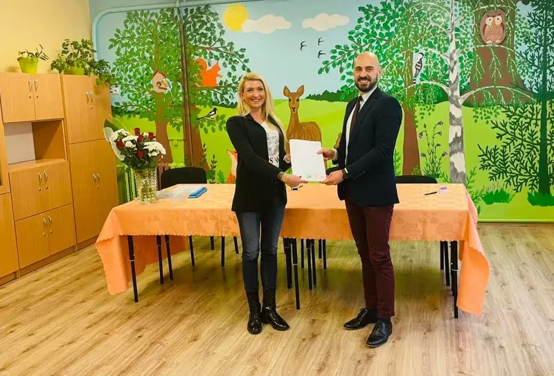 Zdjęcie w formacie poziomym przedstawia kobietę i mężczyznę stojących wewnątrz sali przedszkolnej, trzymających wspólnie biały dokument. Kobieta po lewej stronie ma blond włosy i ubrana jest w czarną marynarkę, białą bluzkę, dżinsy oraz czarne botki. Mężczyzna po prawej stronie ma ogoloną głowę, brodę i nosi granatową marynarkę, białą koszulę z ciemnym krawatem oraz bordowe spodnie. Przed nimi znajduje się stół przykryty pomarańczowym obrusem, na którym stoi wazon z białymi i czerwonymi kwiatami. Ściana w tle jest pomalowana w kolorowy mural przedstawiający las, drzewa, sarnę oraz sowę, co podkreśla edukacyjny charakter wnętrza. Cała kompozycja jest symetryczna i dobrze oświetlona, co czyni ją odpowiednią do publikacji w mediach społecznościowych lub artykułach dotyczących współpracy z placówkami oświatowymi.
