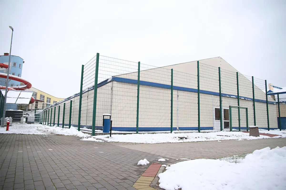 Zdjęcie przedstawia dużą halę namiotową w formacie poziomym, otoczoną wysokim ogrodzeniem z zielonej siatki panelowej. Budynek posiada kremowe ściany z niebieskimi akcentami wykończeniowymi i znajduje się na terenie wyłożonym kostką brukową pokrytą miejscami warstwą śniegu. Po lewej stronie widać fragment niebieskiego budynku z czerwoną, krętą zjeżdżalnią wodną, co sugeruje lokalizację przy kompleksie basenowym. W centralnej części hali widoczne są dwuskrzydłowe drzwi wejściowe, a przed ogrodzeniem stoi niebieski kosz na śmieci. Niebo na zdjęciu jest zachmurzone i jasne, co tworzy równomierne, rozproszone oświetlenie całego obiektu.